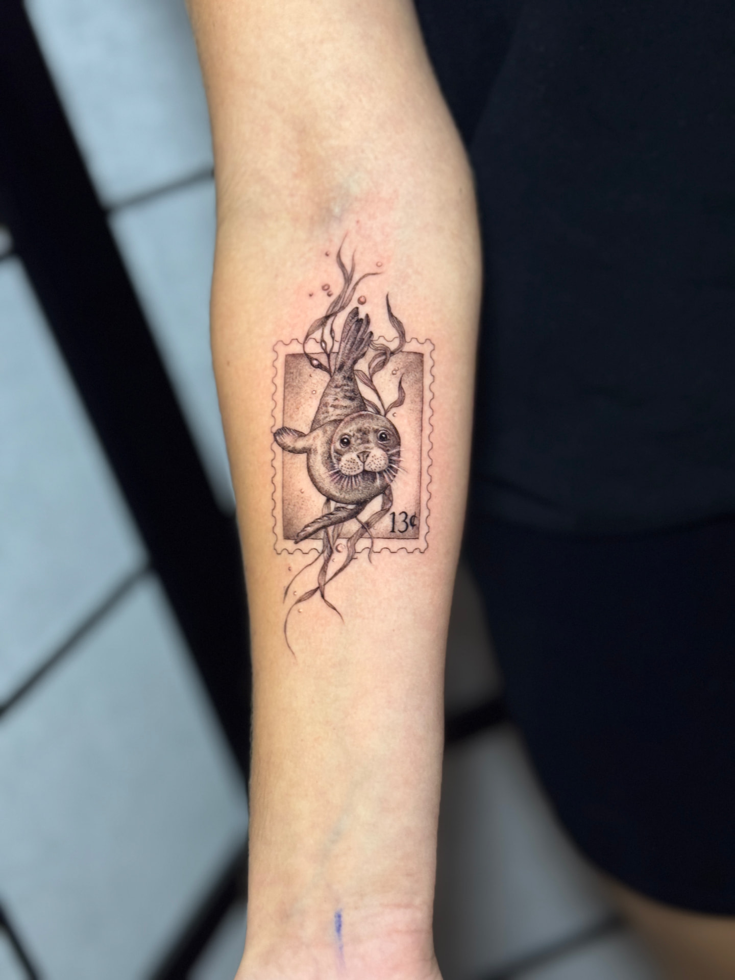 Filigranes Fine-Line-Tattoo mit Robbe als Briefmarke auf dem Unterarm bei Omilian Tattoo bei Kiel